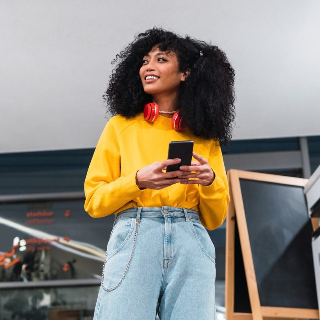 From below of confident positive young ethnic female millennial with dark curly hair in trendy outfit smiling happily and using smartphone while standing in modern office